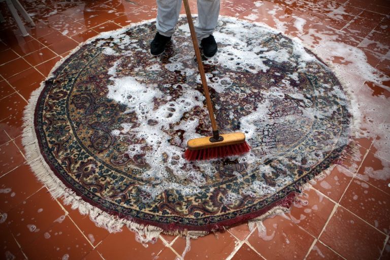 A person using a broom to clean a rug, focusing on removing dirt and debris from the fabric surface.